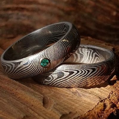 Snubní prsteny z damaškové oceli; vzor damašku napodobuje strukturu dřeva. Dámský prsten je osazen přírodním smaragdem o průměru 2 mm, kámen je upevněn v obrubě z bílého zlata 585/1000.