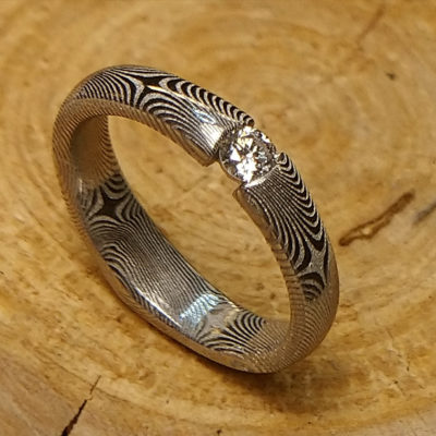  Prsten nevšedního tvaru je vyrobený z vrstvené oceli damasteel a doplněný jiskrným  briliantem o průměru 3 mm. Kámen je zespoda jištěn kroužkem z bílého zlata 585.